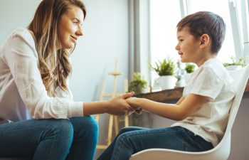 Young boy having therapy with a child psychologist. Smiling woman talking to boy. Friendly young child psychologist talking with little boy suffering from emotional disorder in bright office